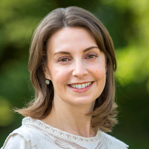 A woman with shoulder-length brown hair, wearing a light-colored top and earrings, smiles warmly at the camera with a blurred green background behind her.