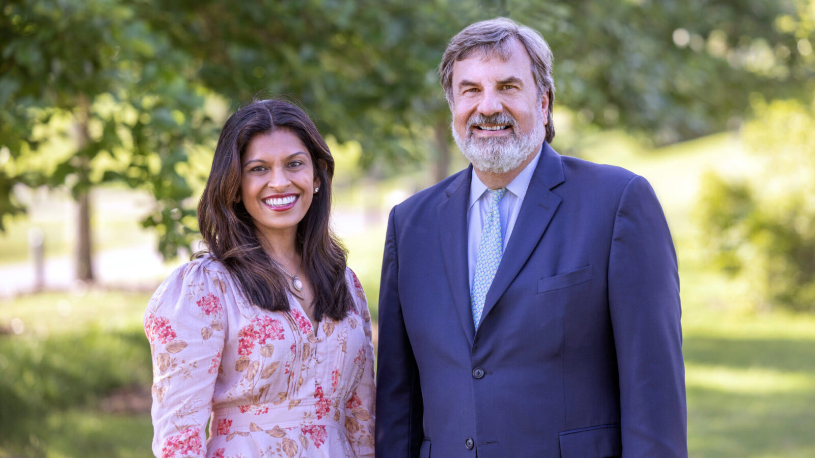 A woman in a floral dress and a man in a dark suit and light blue tie stand side by side outdoors, smiling, with trees and greenery in the background.