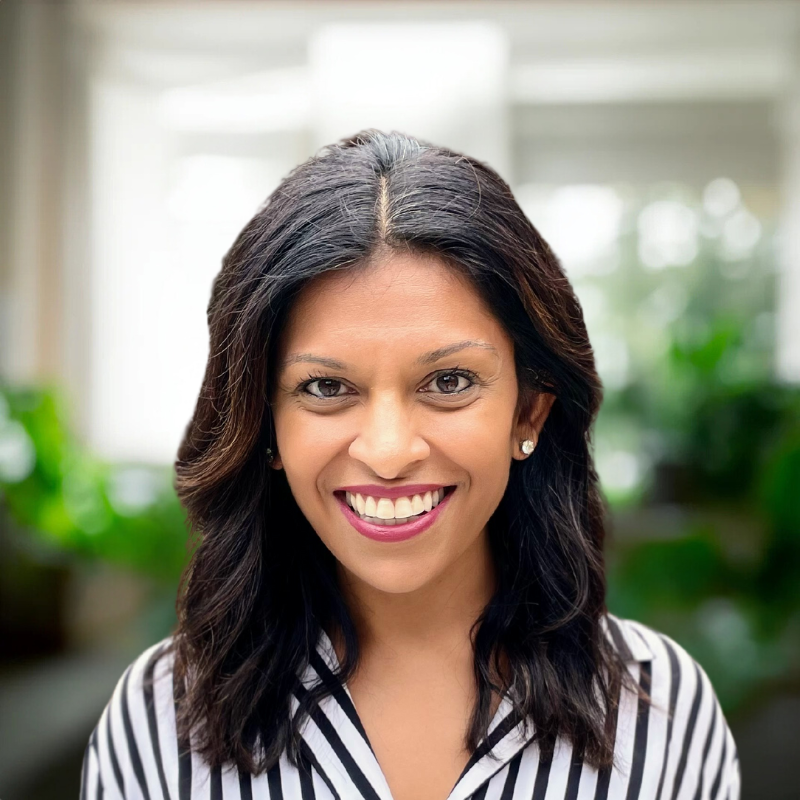 A woman with medium-length dark hair and a bright smile is wearing a black and white striped shirt. She is standing indoors with blurred green plants and large windows in the background.
