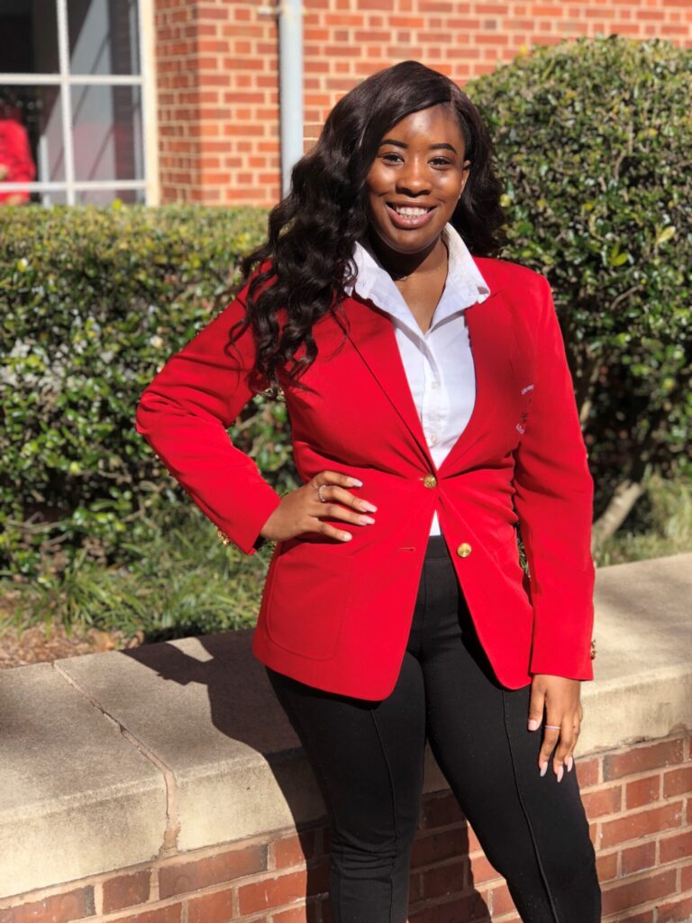 A woman stands outside in sunlight, smiling confidently in a red blazer over a white shirt and black pants—perhaps celebrating recent scholarships. She has long, wavy hair and rests one hand on her hip. Bushes and a brick building are visible in the background.