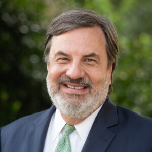 A middle-aged man with gray hair and a beard, wearing a dark suit, white shirt, and light green tie, smiling outdoors with a blurred green background.