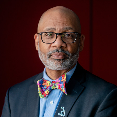 A man with glasses, a gray beard, and a bald head wears a suit, a blue shirt, and a colorful puzzle-patterned bow tie. He stands against a dark red background.