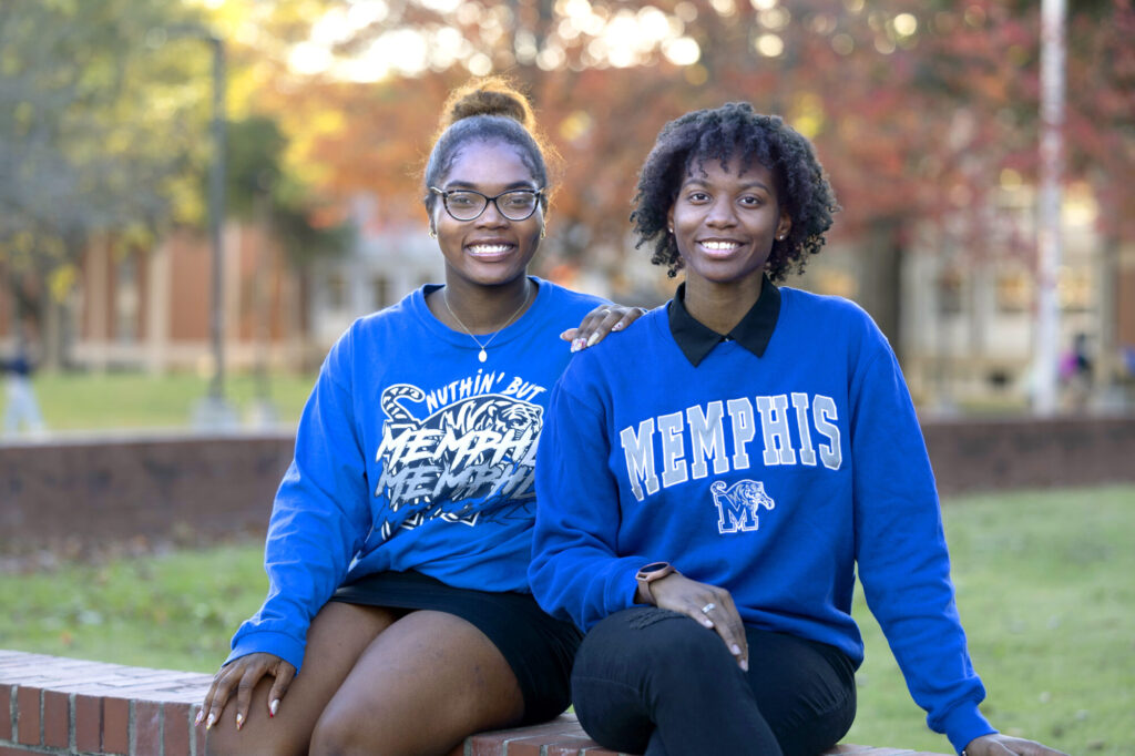Two young women sit outdoors on a brick ledge, smiling at the camera in blue University of Memphis sweatshirts. Autumn trees and a campus building are visible, reflecting the possibilities that scholarships bring to student life.