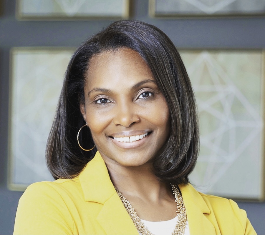 A woman with straight, shoulder-length hair smiles at the camera. She is wearing a yellow blazer, white top, gold hoop earrings, and a gold necklace. The background features geometric artwork in muted colors.