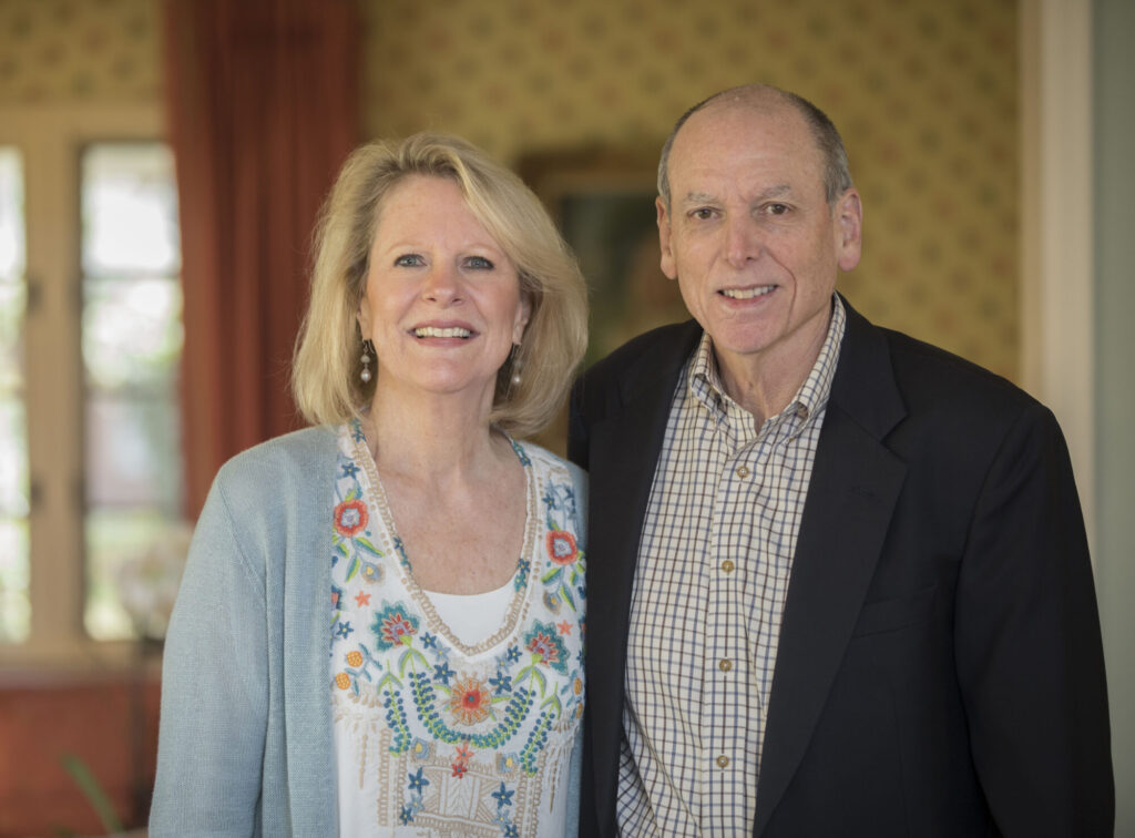 A smiling middle-aged couple stands indoors. The woman has shoulder-length blonde hair, wearing a light blue cardigan and a patterned blouse. The man has short gray hair and wears a black blazer over a checked shirt.