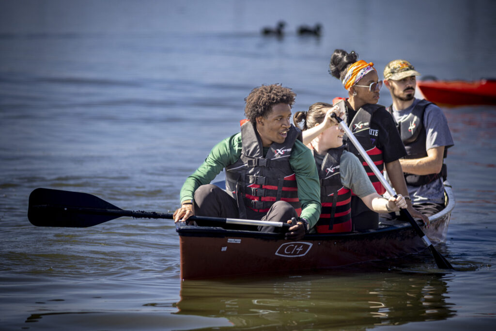 Four people wearing life jackets paddle together in a brown canoe on calm water, embodying teamwork and shared values, with blurred ducks in the background. All appear focused and engaged in the activity.