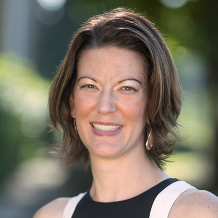 A woman with short brown hair smiles at the camera. She is wearing a sleeveless black and white top and hoop earrings. The background is blurred greenery, suggesting an outdoor setting.