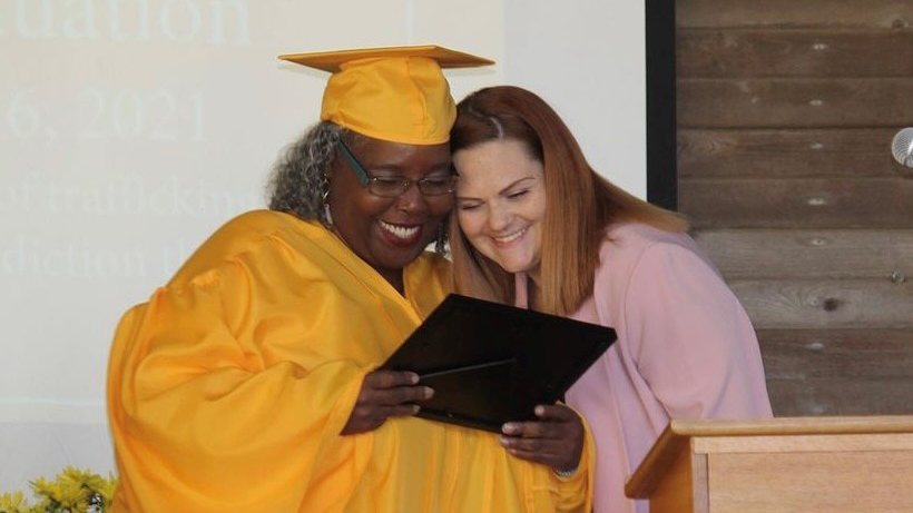Two women smile and look at a certificate together. One, wearing a bright yellow graduation cap and gown, celebrates her achievement of earning scholarships, while the other in a pink outfit stands supportively beside her at the podium.