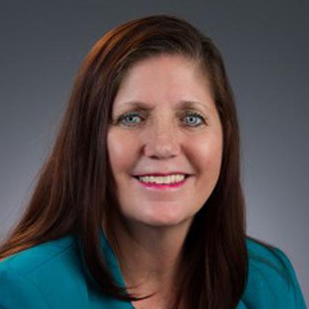 Alisa Haushalter, a woman with long brown hair and blue eyes, wears a teal blazer as she smiles at the camera against a plain gray background.
