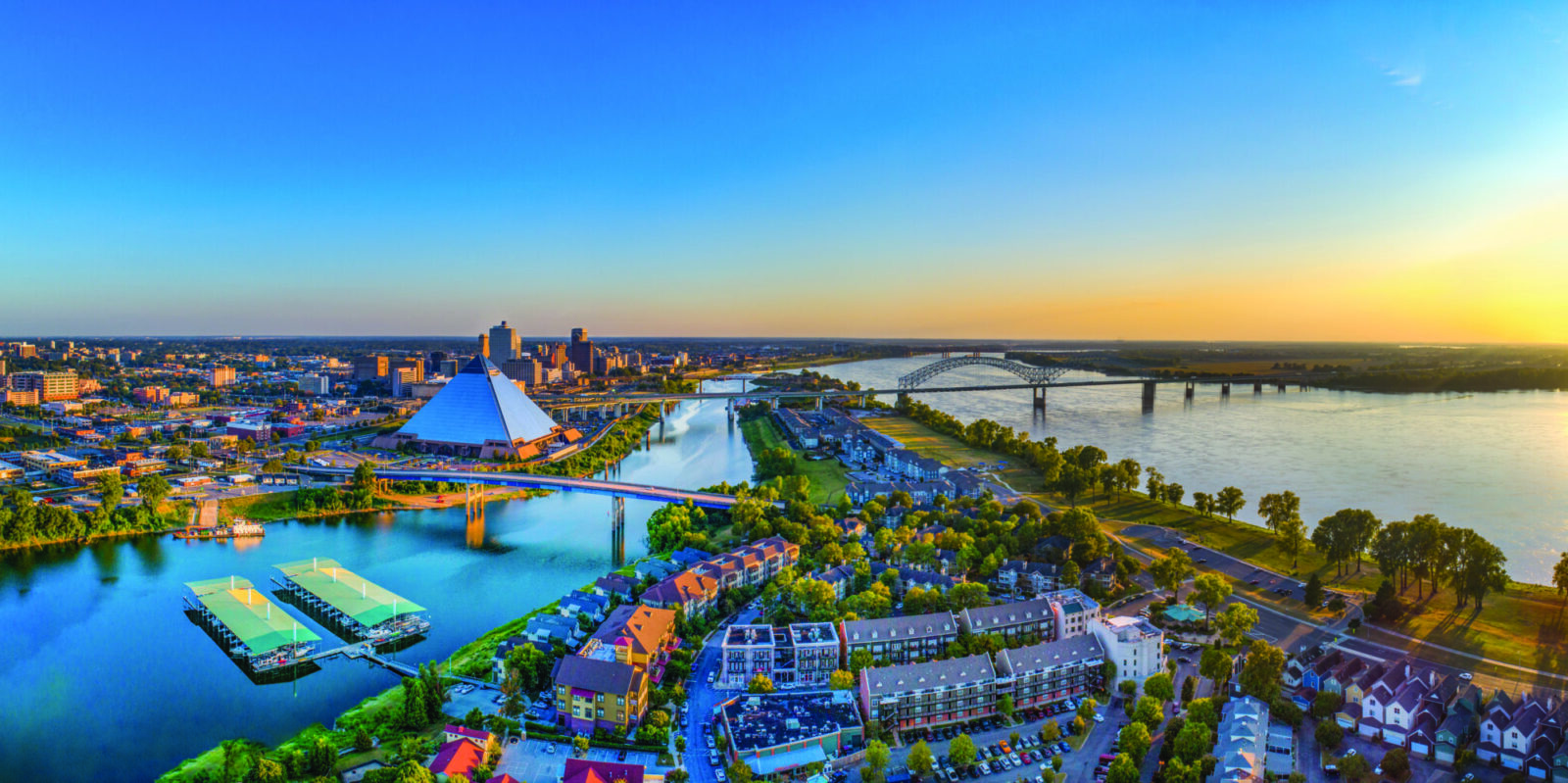 Aerial view of a cityscape at sunset with a river, bridges, a glass pyramid building, high-rise downtown buildings, green areas, and residential neighborhoods in the foreground.