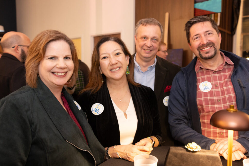 Four adults, three women and one man, pose and smile together at an indoor event. They are dressed in semi-formal attire and all wear round stickers labeled 365. Warm lighting creates a welcoming atmosphere.