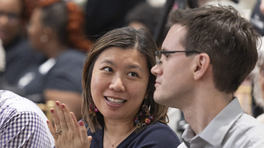 A woman smiling and clapping while discussing recent grants with a man in glasses at an indoor event, with other people blurred in the background.