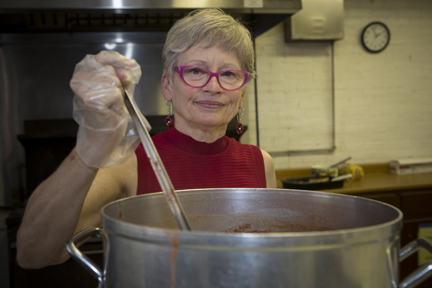 An older woman wearing glasses and a red sleeveless top stirs a large pot of food in a kitchen, using a ladle and wearing a plastic glove. A clock and kitchen equipment are visible in the background.