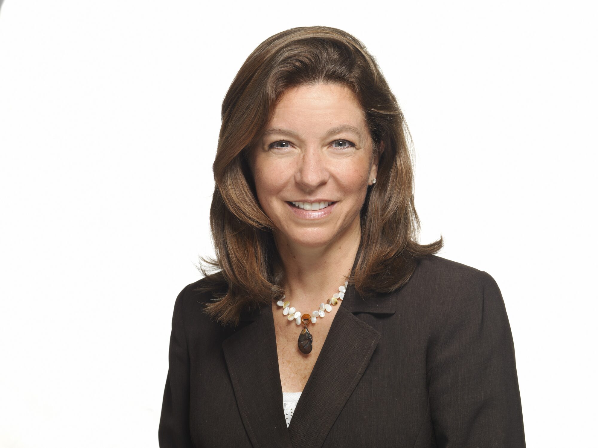 A woman with long brown hair, wearing a dark blazer and a beaded necklace, smiles at the camera against a plain white background.