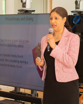 A woman in a pink blazer speaks into a microphone while standing in front of a screen displaying a presentation titled Philanthropy and Giving, emphasizing the importance of continuing education. She holds a small device in her other hand.