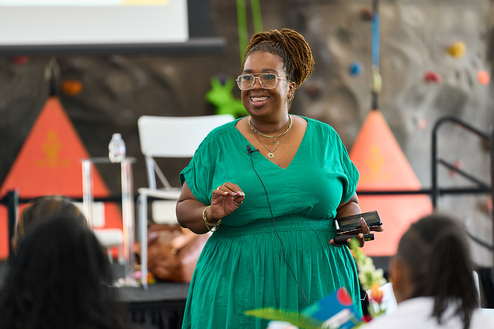 A woman in a green dress smiles while speaking to an audience. She holds a phone and gestures with her hand, standing in a well-lit room with orange and gray backgrounds. People are seated and listening in the foreground.