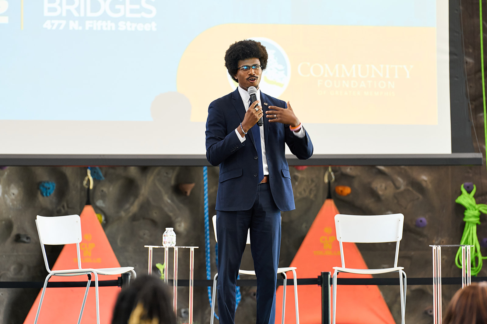 A man in a blue suit speaks into a microphone on stage in front of a screen displaying the Community Foundation logo, with two empty white chairs beside him and an indoor climbing wall in the background.