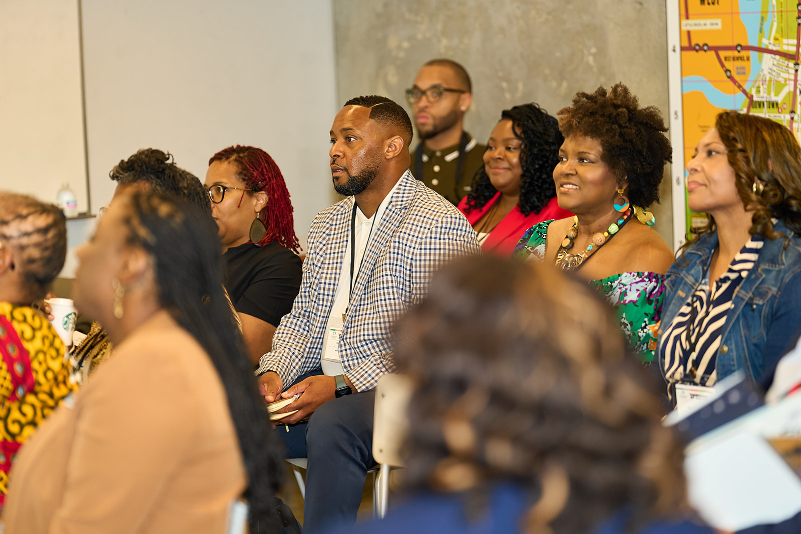 A group of adults sit attentively in a brightly lit room, listening to a speaker. Most are dressed in business or business-casual attire. A colorful map is visible on the wall in the background.