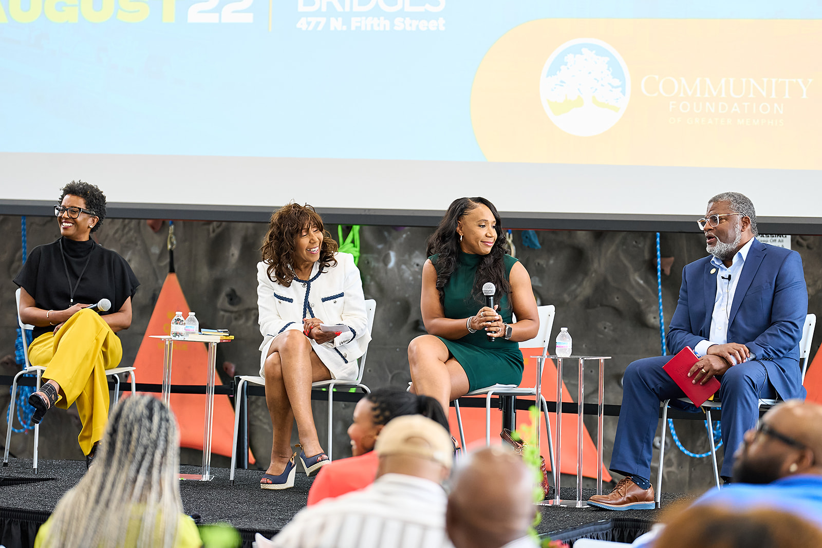 Four people sit on a stage panel, smiling and talking into microphones. They are seated in front of a large screen displaying event details. Audience members are visible in the foreground.
