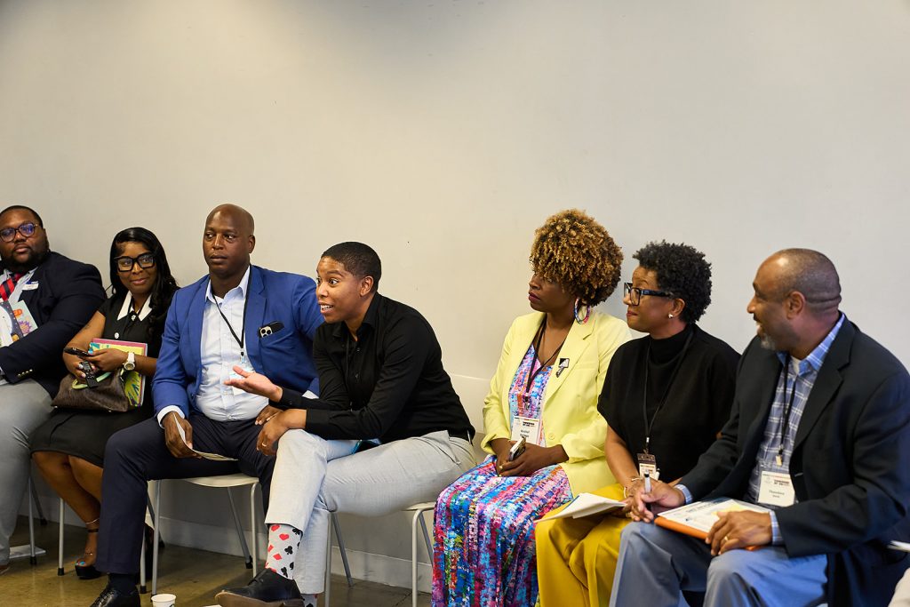 A diverse group of seven adults, including a Black Philanthropic Advisor, sit in a row engaged in discussion. One speaks animatedly while others listen attentively, holding notebooks and pens against a plain gray wall background.