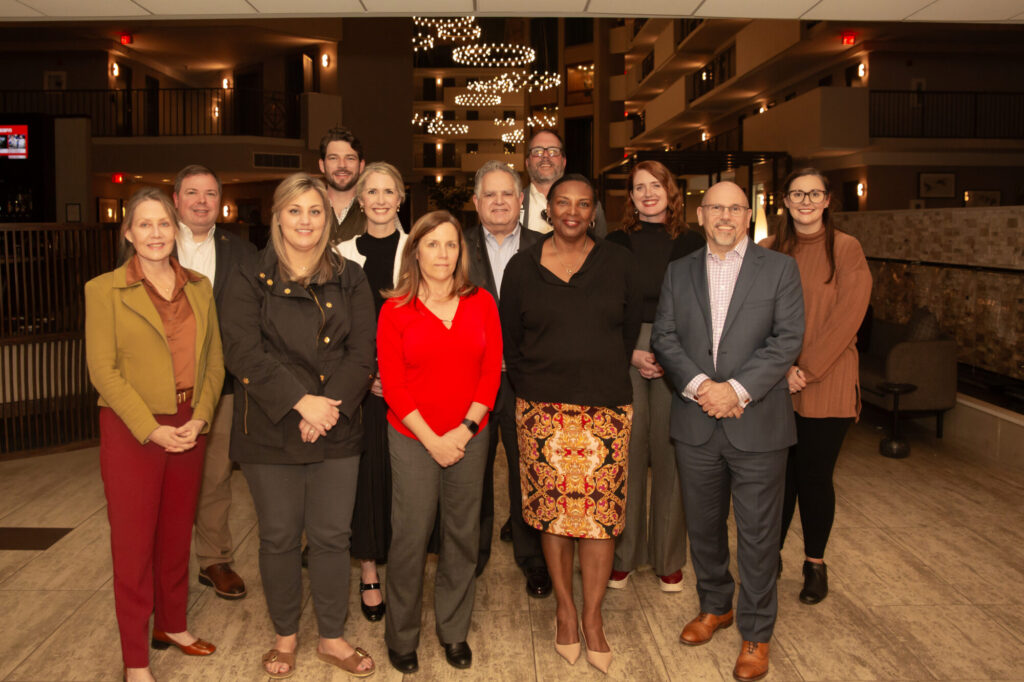 A group of twelve professionally dressed adults, including a Chartered Advisor in Philanthropy (CAP designation), stands together indoors, posing and smiling for a group photo in a warmly lit, modern lobby with decorative lighting in the background.