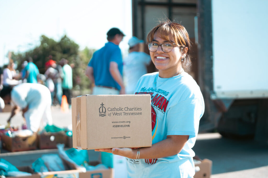 A smiling woman wearing glasses and a light blue shirt holds a box labeled Catholic Charities of West Tennessee at an outdoor donation event, where volunteers distribute food, scholarships, and grants to support the local community.