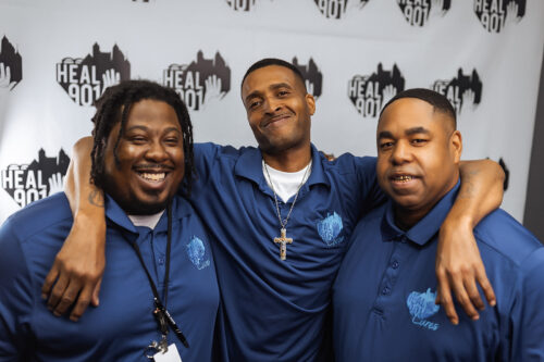 Three men smiling and standing together, all wearing matching blue shirts with “Heal 901” logos, in front of a backdrop featuring the same logo. The group recently celebrated receiving recent grants to support their community initiatives.