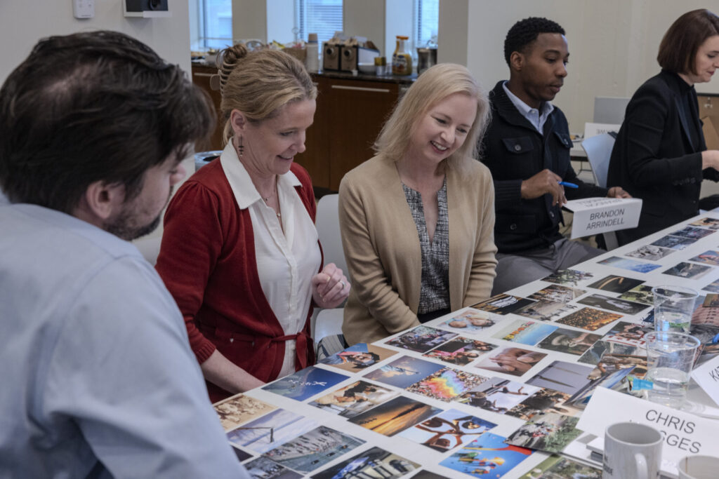 Four people sit around a table covered with various photographs, looking at them and smiling. Office supplies and coffee are in the background, as name cards suggest this gathering is part of a Next Gen philanthropic initiative.