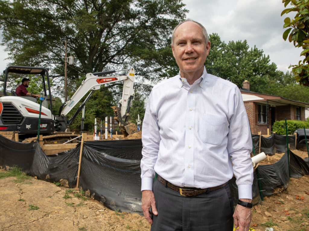 A man in a white dress shirt stands in front of a construction site with a small excavator and black silt fencing, next to a brick house surrounded by trees on a cloudy day.