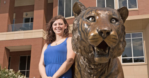 A woman in a blue dress stands smiling next to a large bronze tiger statue in front of a modern brick building, celebrating her achievements and scholarships.