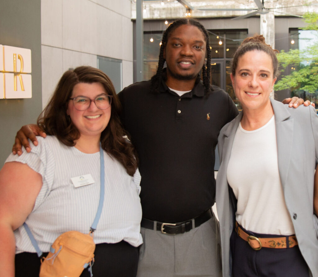 Three people stand smiling together outdoors near a modern building. A woman with glasses and a tan bag stands on the left, a man in a black shirt is in the center, and a woman in a gray blazer is on the right.