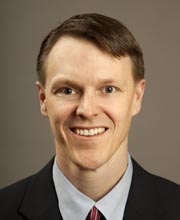 Lance Hollingsworth, CFP, with short light brown hair, fair skin, and blue eyes, is smiling in a dark suit jacket, light blue dress shirt, and red-striped tie against a plain beige background.