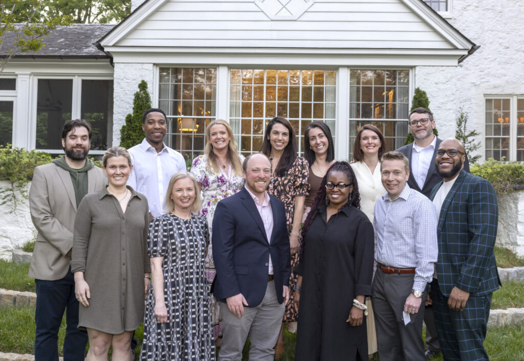 A group of thirteen adults stands smiling in two rows in front of a white house with large windows and greenery. Most are wearing business casual attire, gathered for a Next Gen Philanthropic Initiative outdoors in the early evening.