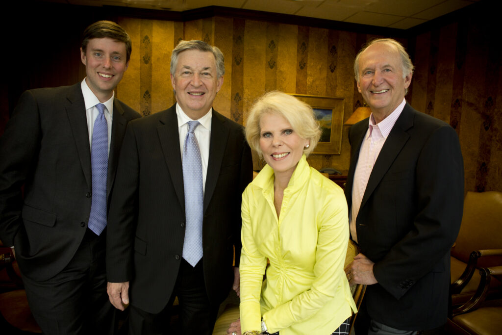 Four professionally dressed adults, three men in suits and ties and one woman in a yellow blouse, smile and pose together in an office with warm lighting and patterned walls, reflecting their shared commitment to philanthropic education.