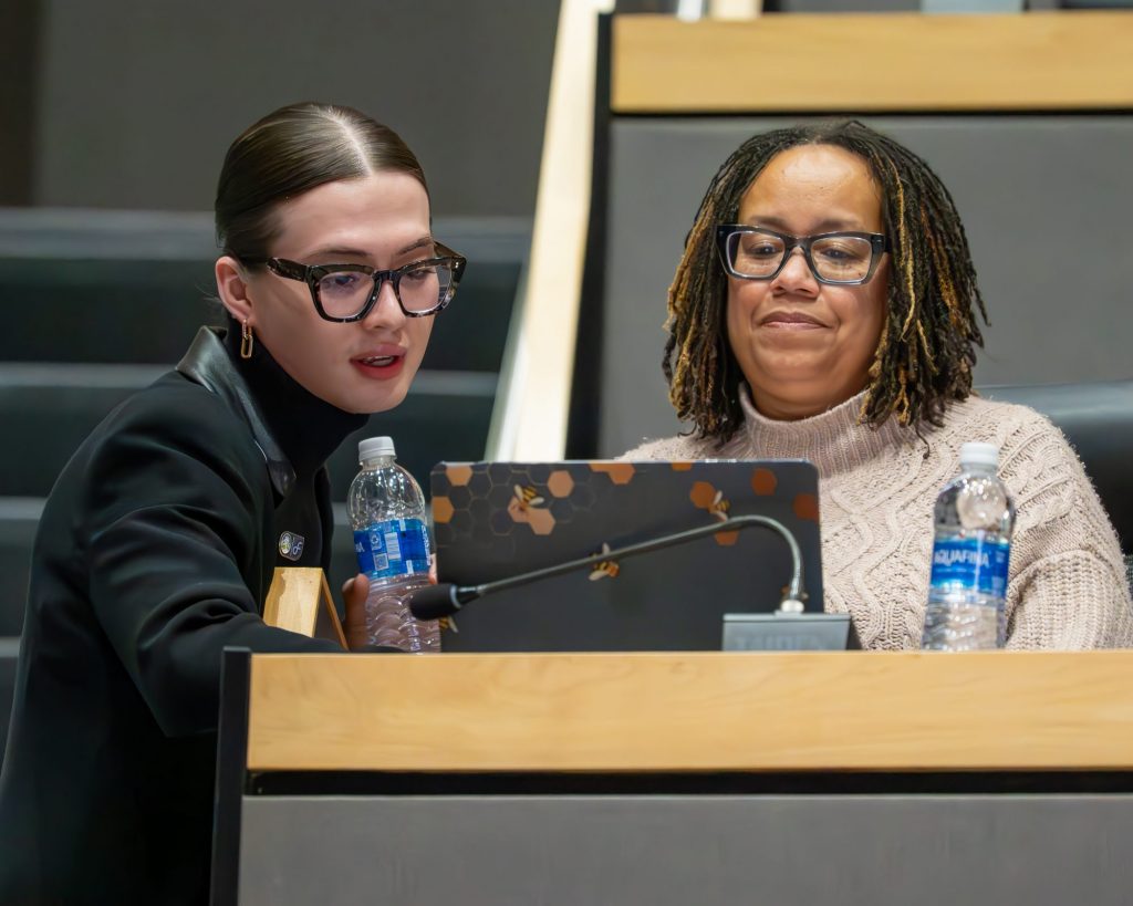 Two women sit together at a desk with a laptop and water bottles, exploring resources for their project. One with glasses and braided hair looks at the screen, while the other, also in glasses, speaks and holds a water bottle during their trainings.
