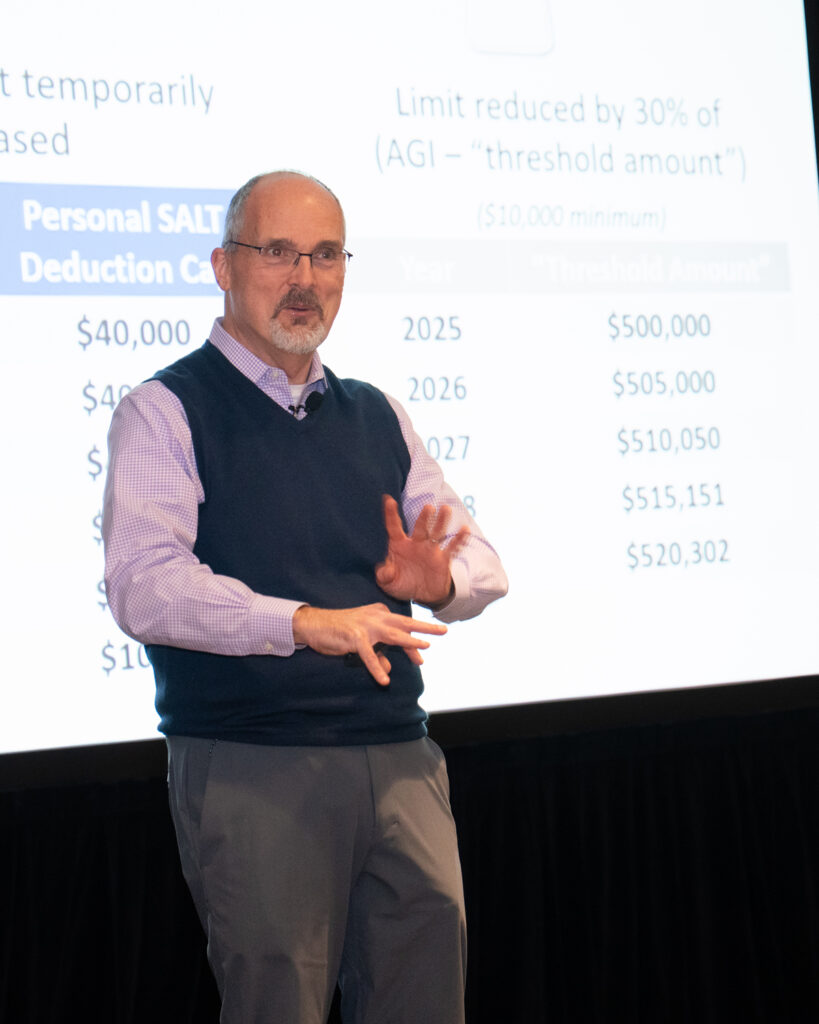 A middle-aged man wearing glasses, a light purple shirt, and a navy sweater vest presents financial data and charts on a projected slide, gesturing as he explains the information during a continuing education session.