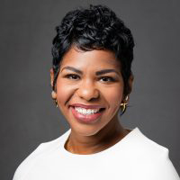 Dr. Shanea McKinney, a woman with short, curly black hair, smiles at the camera. She is wearing gold hoop earrings and a white top, with a dark gray background behind her.