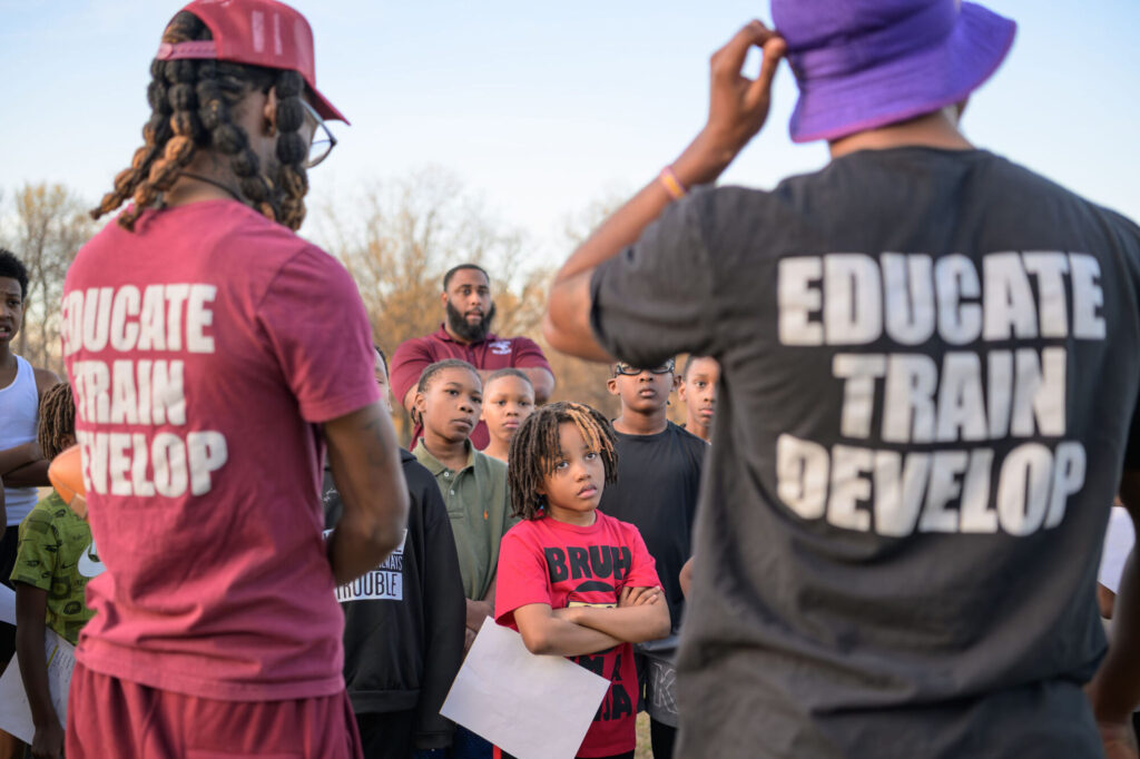 A group of children stand outdoors listening to two adults whose shirts read “EDUCATE TRAIN DEVELOP.” The children look attentive, with trees and a clear sky in the background.