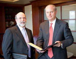 Two professional advisors in suits stand in an office, one holding a folder and the other a notepad. Bookshelves and wood paneling are visible in the background. Both men have neutral expressions.