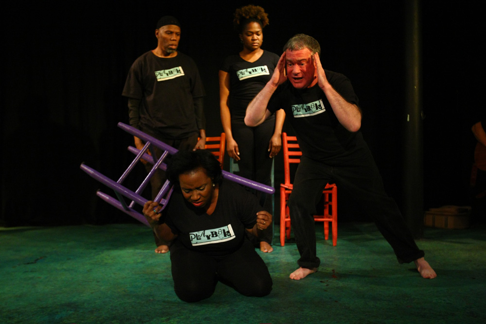 Four actors perform on stage. One woman kneels, holding a purple chair, looking distressed—a scene reflecting debates about grantmaking priorities. A man beside her holds his head dramatically; two others stand behind, all in black shirts with white text.