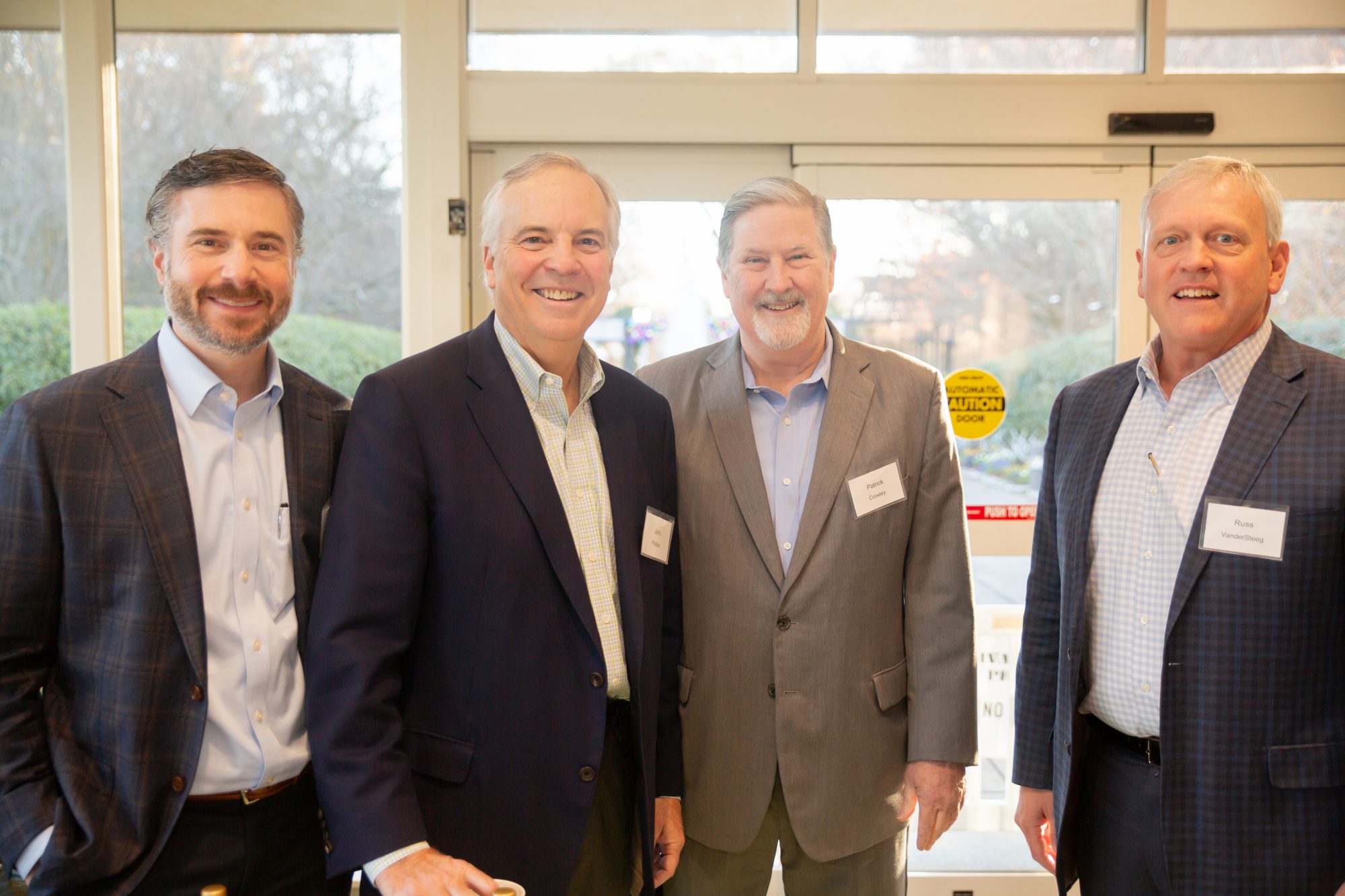 Four men in business attire stand together indoors, smiling at the camera. They are wearing name tags and suits. A glass door and outdoor greenery are visible in the background.