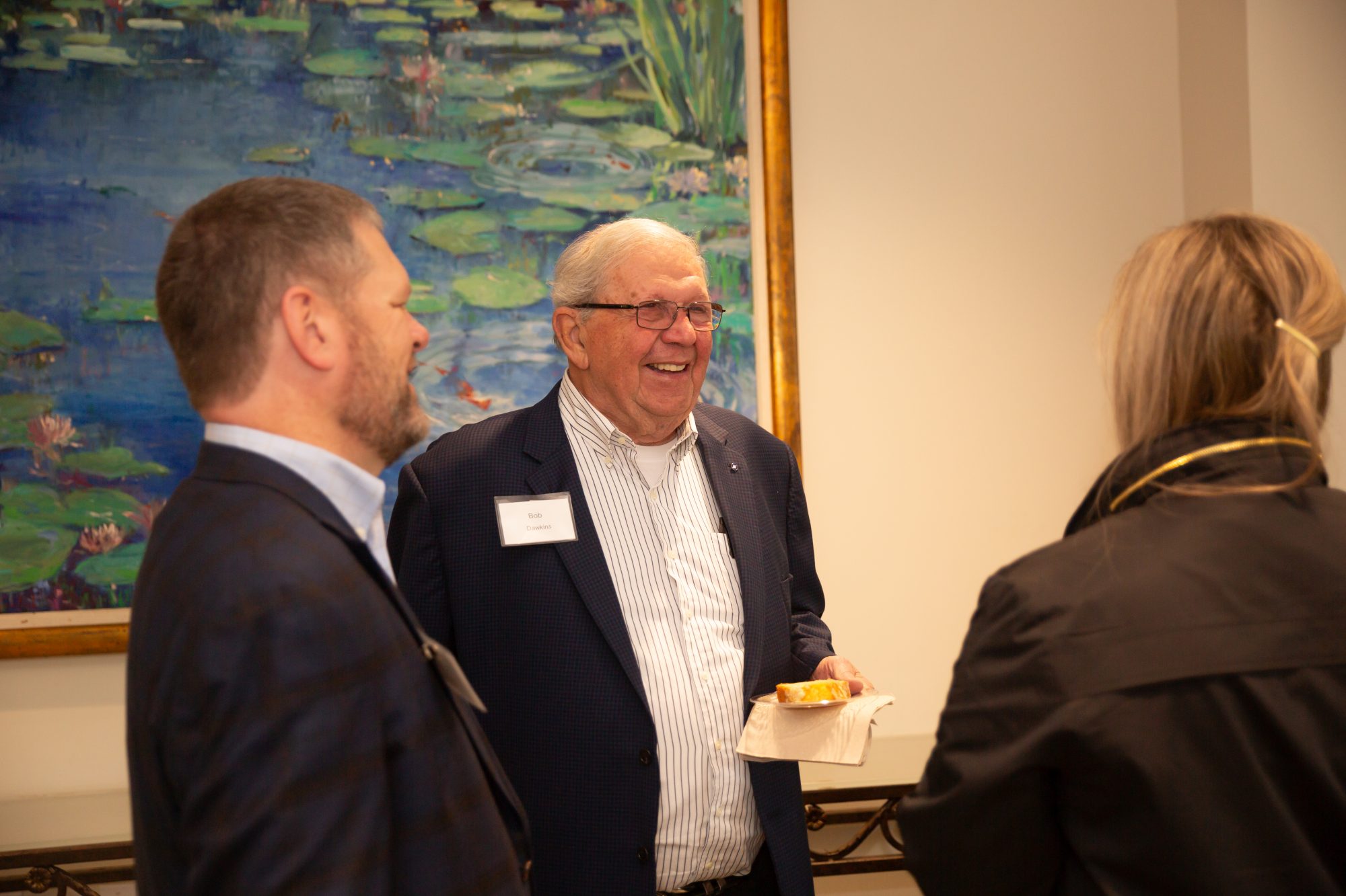 Three people are talking and smiling at an indoor event. One older man holds a plate with food. A colorful painting of water lilies hangs on the wall behind them.
