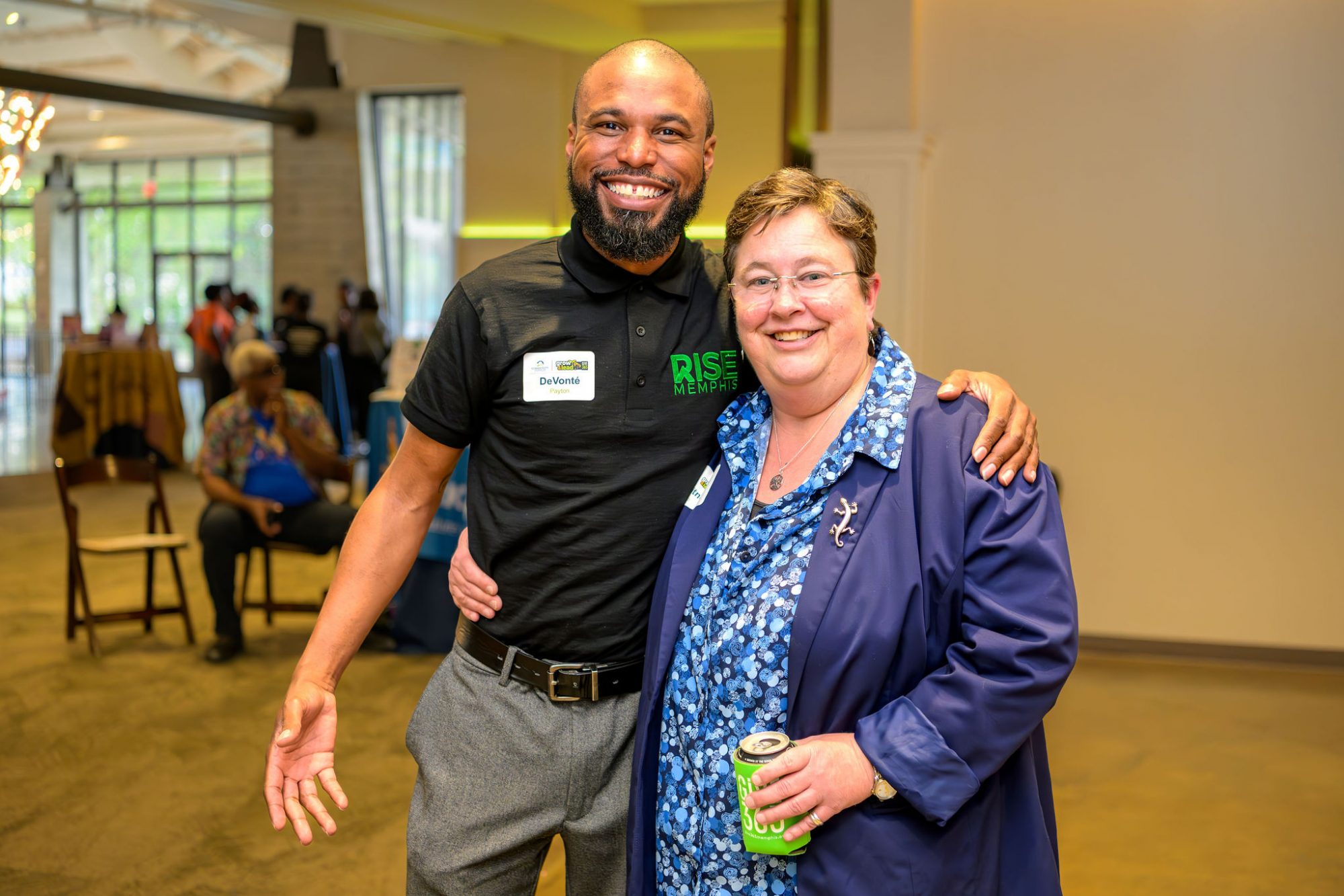 A man and woman smiling and posing together indoors at an event; the man wears a black polo and the woman wears glasses with a blue jacket and holds a can. Other people are visible in the background.