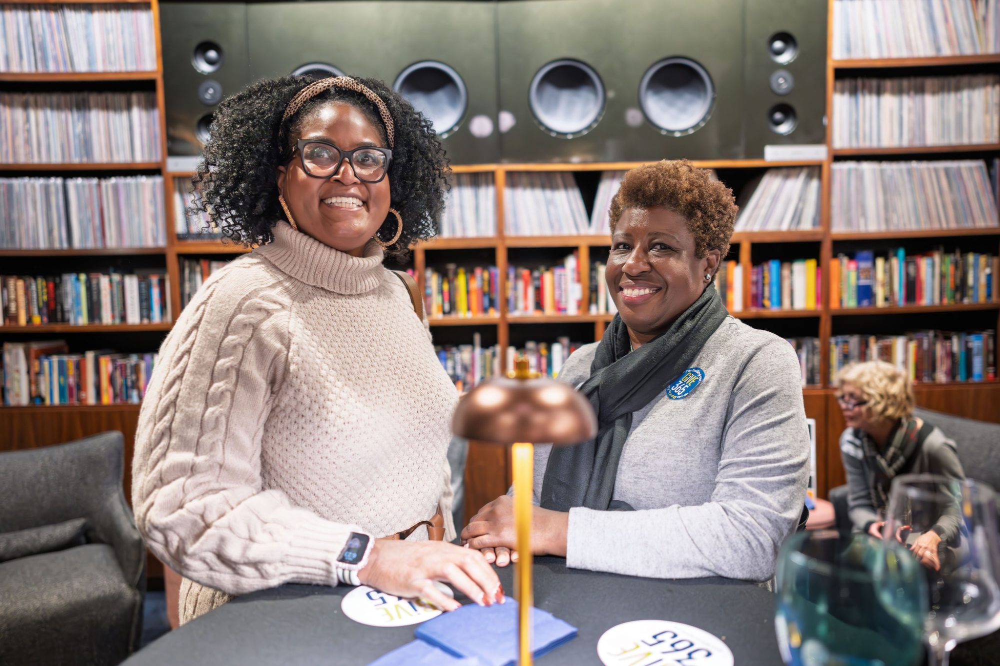 Two women smiling stand at a round table in a cozy library or lounge, lined with books and records. One wears a light sweater and glasses, the other a blazer. A seated person reads in the background.