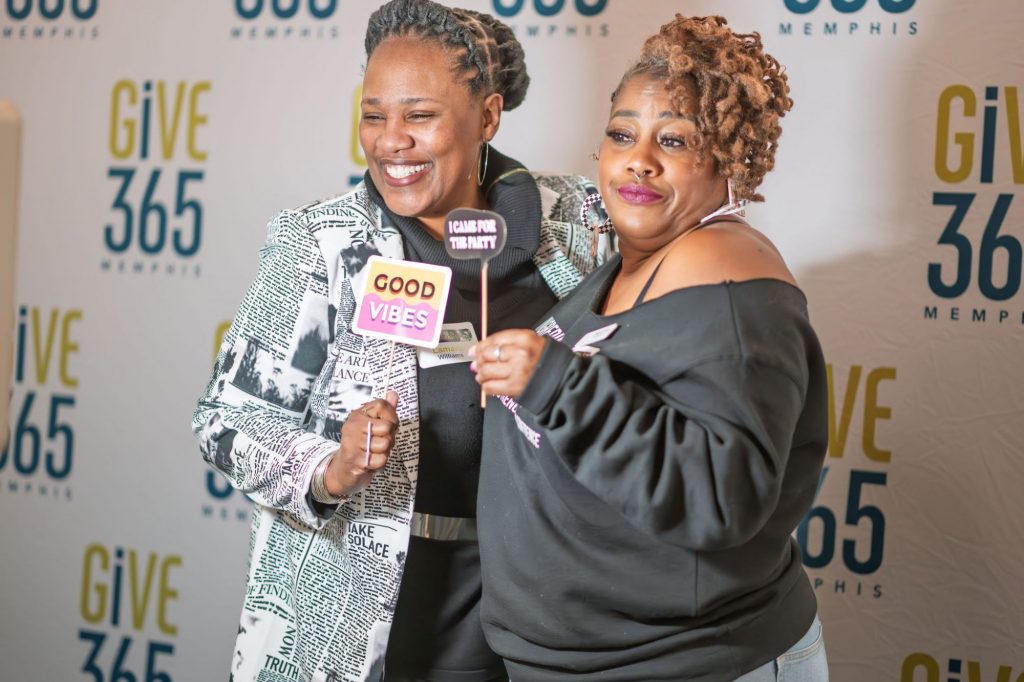 Two smiling women pose together at a Give 365 Memphis event, holding playful signs that read “GOOD VIBES” and “I CAN PROVE IT WITH A T-SHIRT,” standing in front of a branded photo backdrop.
