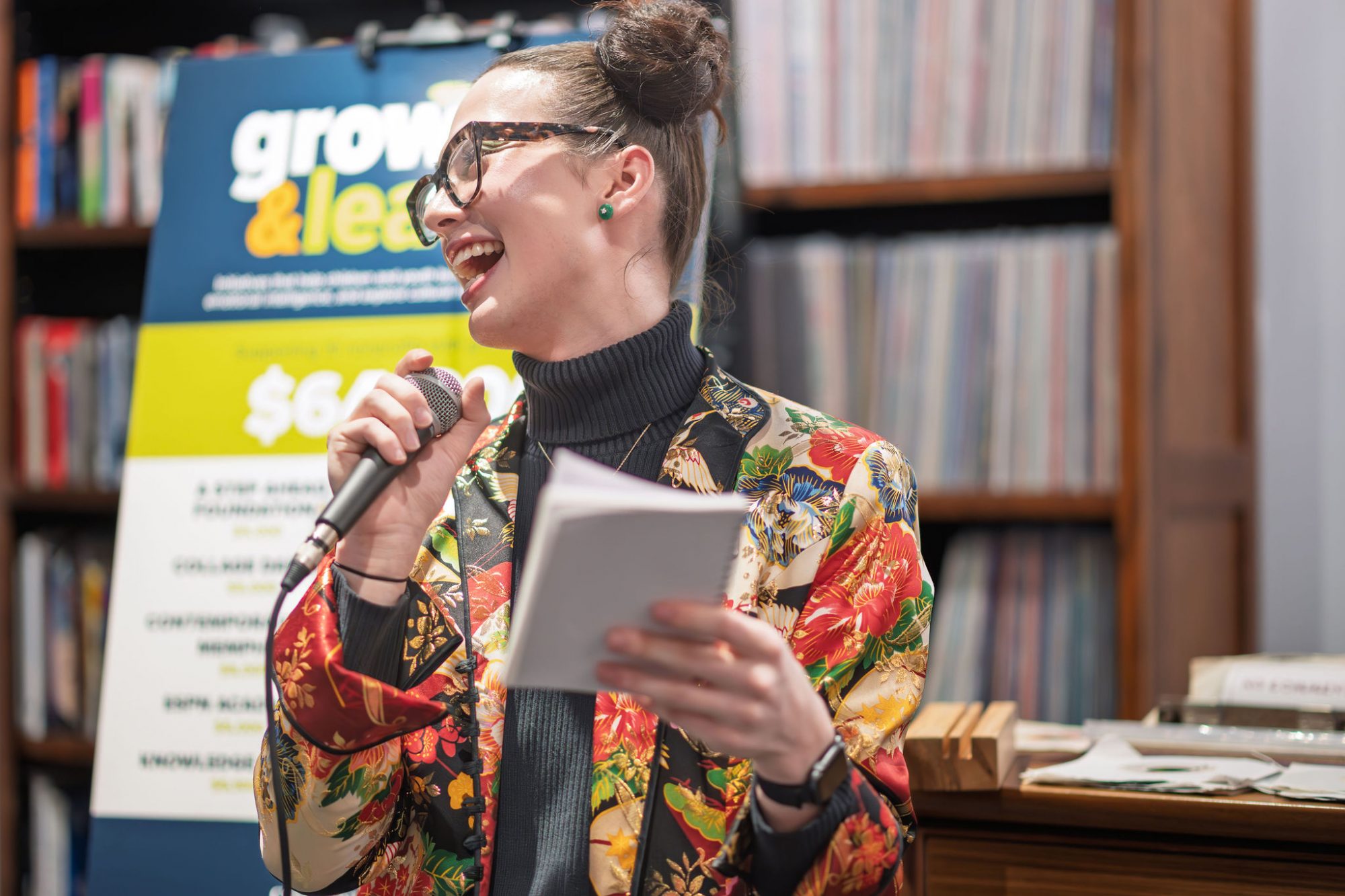 A person with glasses and a floral jacket smiles while speaking into a microphone and holding a notebook, standing in front of a sign and bookshelves.