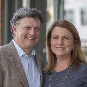 Kevin Brewer in a plaid blazer and Cindy Brewer in a blue top stand close together, smiling at the camera in an outdoor setting.