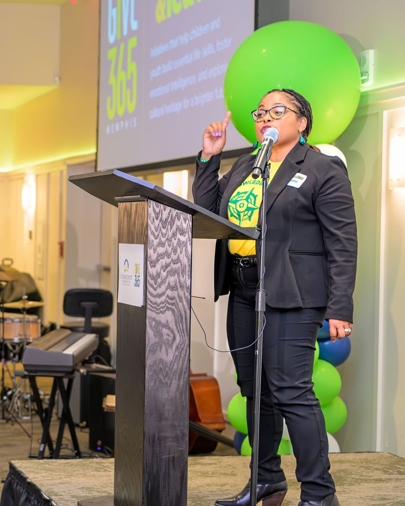 A woman wearing glasses and a suit speaks at a podium on a stage decorated with green and blue balloons, with a projector screen and musical instruments visible in the background.