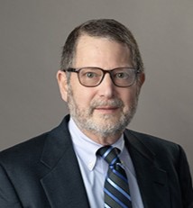 A middle-aged man with short brown hair, a beard, and glasses wears a dark suit, light blue shirt, and striped tie, posing against a plain gray background—perfect for promoting your next Planned Giving seminar.