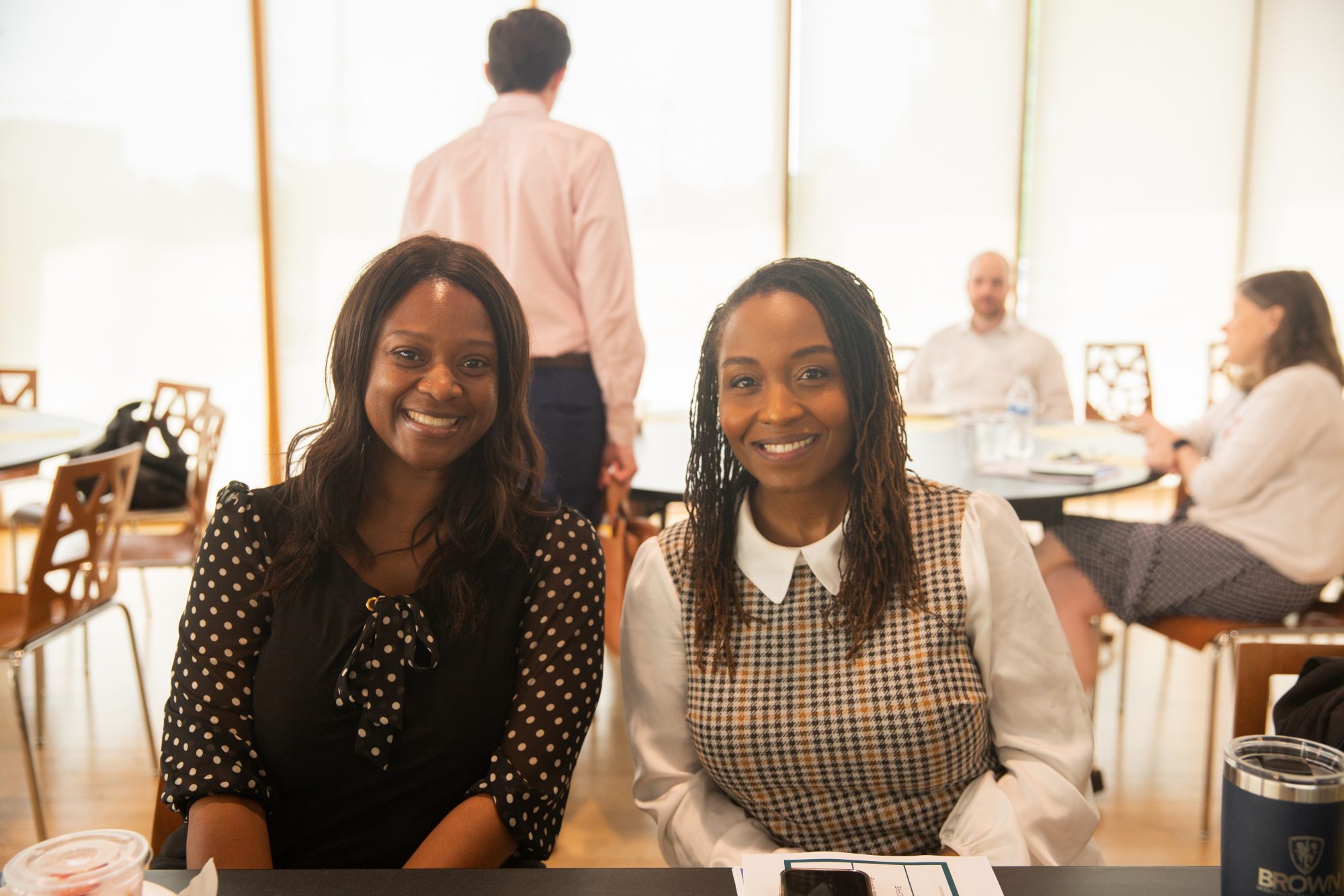 Two women are smiling at a table in a bright, modern room with other people sitting and talking in the background. Papers, drinks, and a phone are visible on the table in front of them.